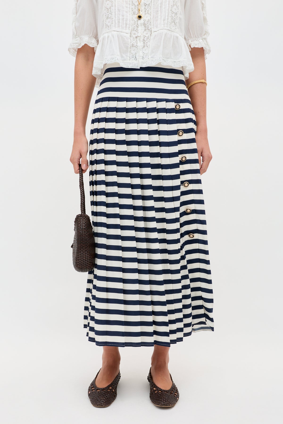 A person wears the Una - Lea Stripe Navy pleated skirt, a white lace blouse, and holds a woven brown bag with matching flats. The photo is cropped just below the shoulders.