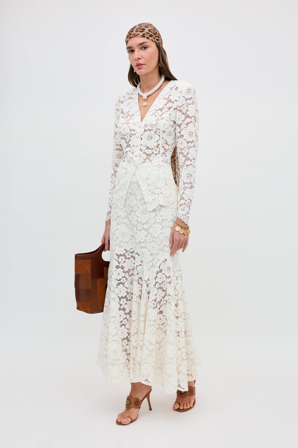 A woman stands against a plain background wearing the Jada - Cream long lace dress with long sleeves, tan heels, a brown patterned headscarf, layered necklaces, gold jewelry, and carrying a brown handbag.