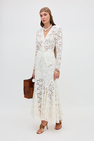 A woman stands against a plain background wearing the Jada - Cream long lace dress with long sleeves, tan heels, a brown patterned headscarf, layered necklaces, gold jewelry, and carrying a brown handbag.