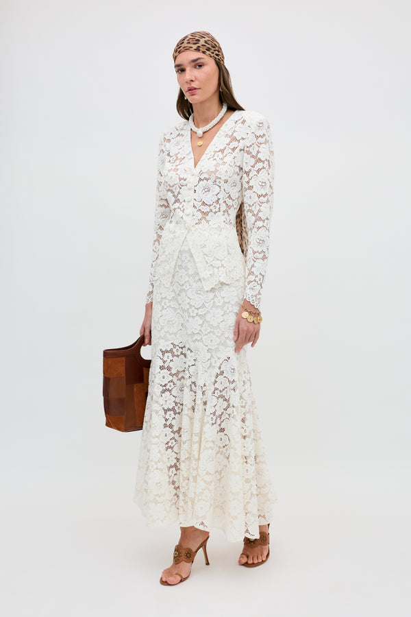 A woman stands against a plain background wearing the Jada - Cream long lace dress with long sleeves, tan heels, a brown patterned headscarf, layered necklaces, gold jewelry, and carrying a brown handbag.
