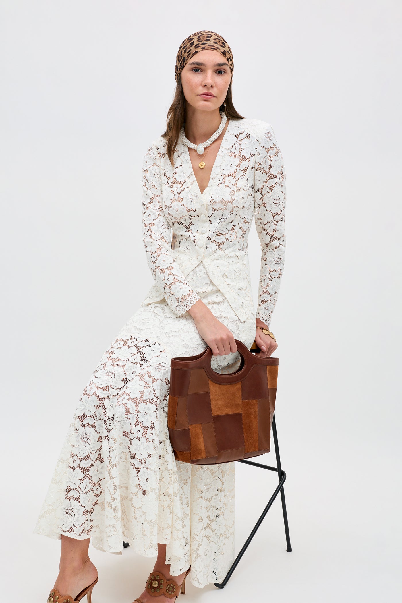 Wearing the Jada - Cream, a long white lace dress, a woman sits on a black chair with a brown patchwork bag, leopard print headscarf, gold charm necklace, and brown heels against a plain light background.