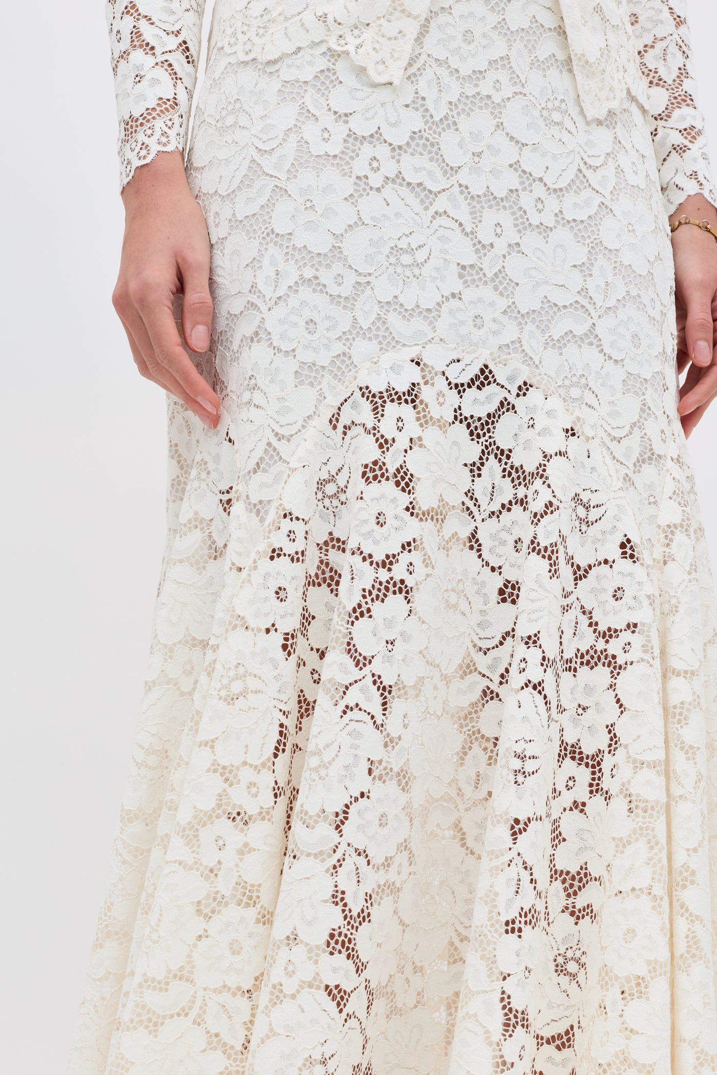 A close-up of a person wearing the Jada - Cream dress, featuring long white lace with floral patterns, shown from the waist to the knees and one hand resting at their side.