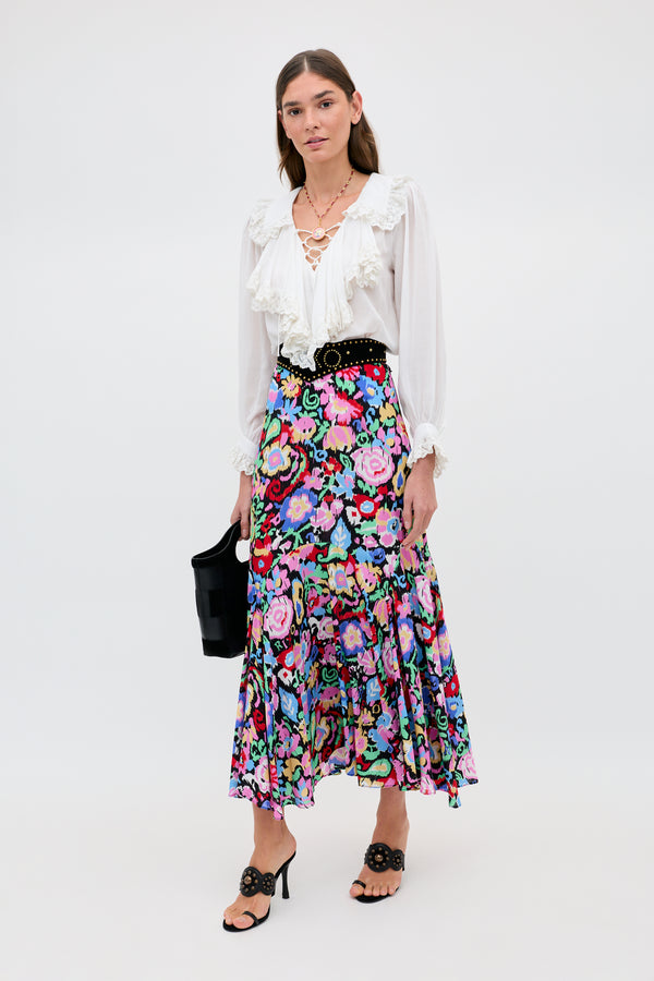 A woman poses against a plain background wearing the Libbie - Ikat Bloom Charcoal floral midi skirt, a white ruffled blouse, black sandals with a matching belt, layered necklaces, and carrying a black handbag.