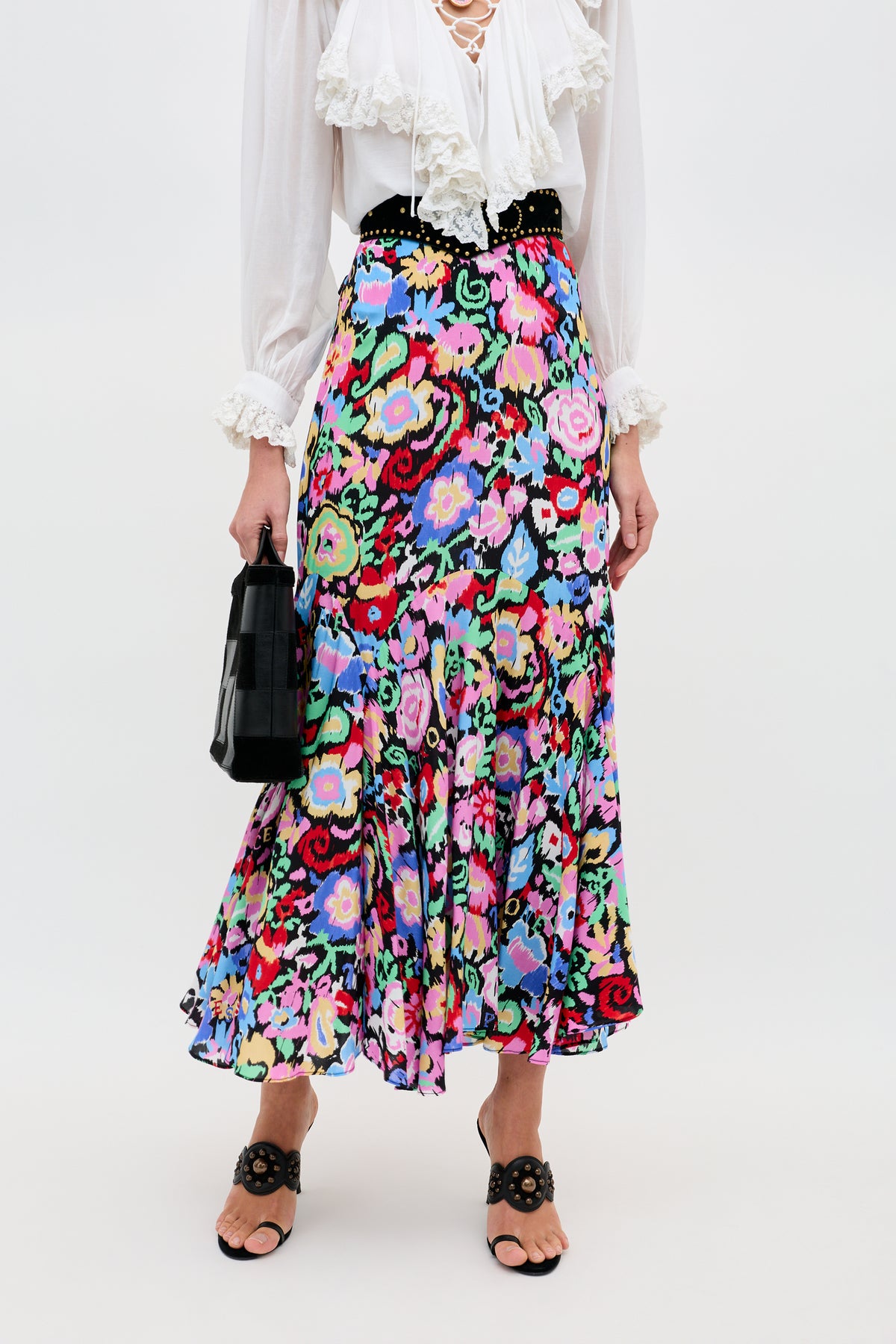 A person stands against a plain white background wearing the Libbie - Ikat Bloom Charcoal floral midi skirt, a white ruffled blouse, a black handbag, and black open-toe sandals with floral designs.