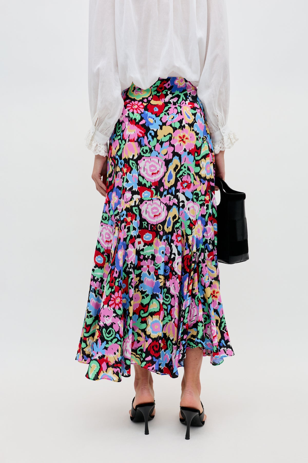 A person stands facing away, wearing the Libbie - Ikat Bloom Charcoal colorful floral midi skirt, a white long-sleeve blouse, black high-heeled sandals, and holding a small black bag against a plain background.