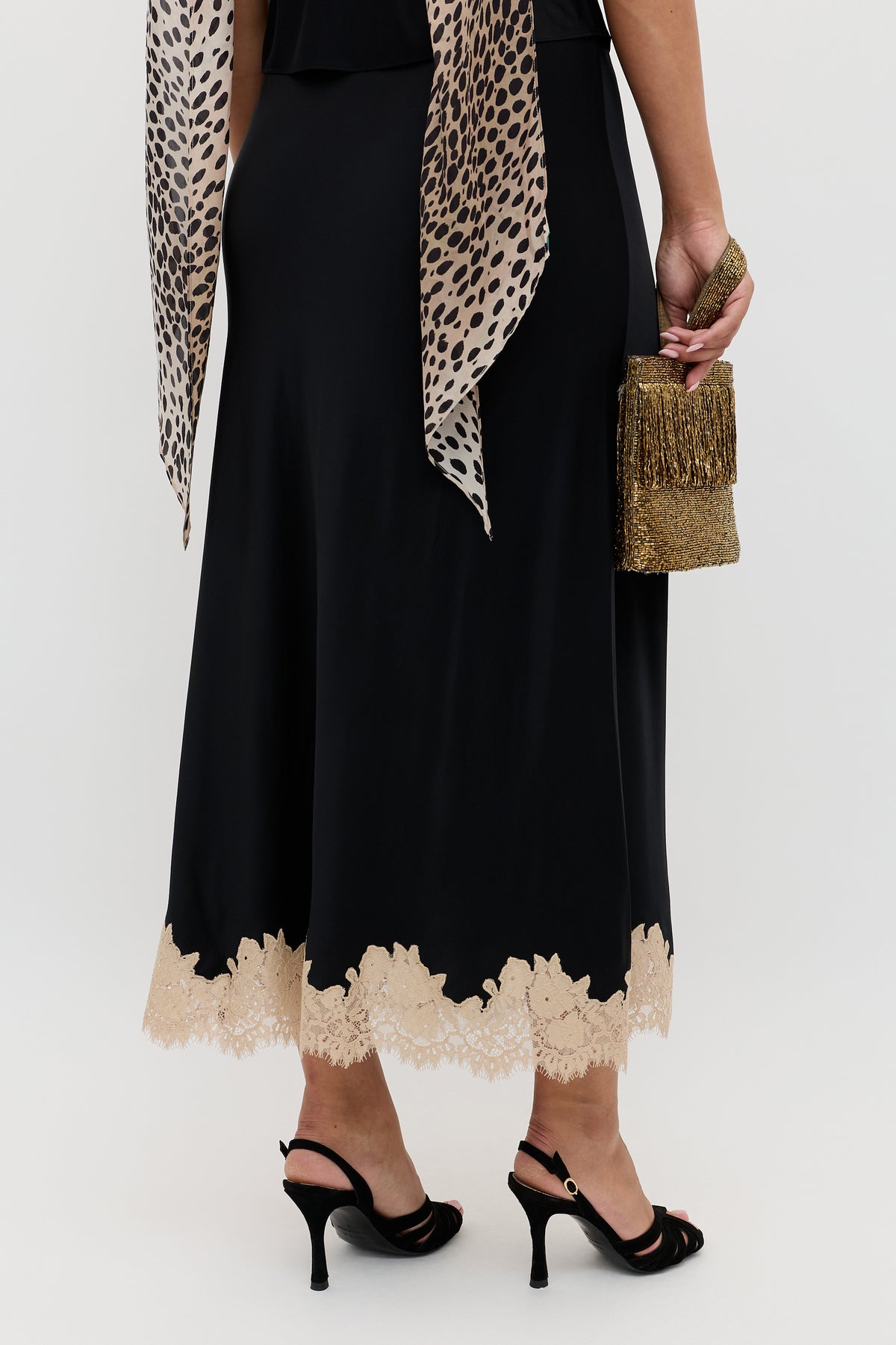 A person wearing the Matilde - Black skirt with beige lace trim, paired with black heels and holding a gold fringed clutch. A leopard-print scarf drapes from their shoulders, all set against a plain white background.