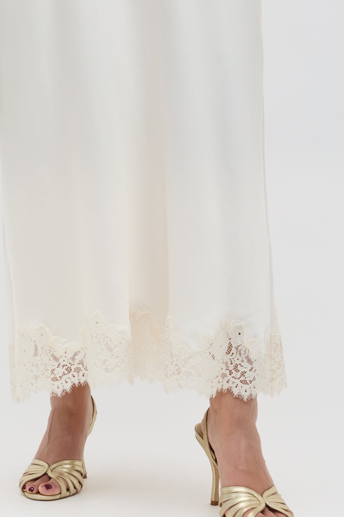 A close-up of a person wearing the Matilde - Pearl long white satin skirt with lace trim, paired with gold open-toe heels for an elegant bridal look against a plain white background.