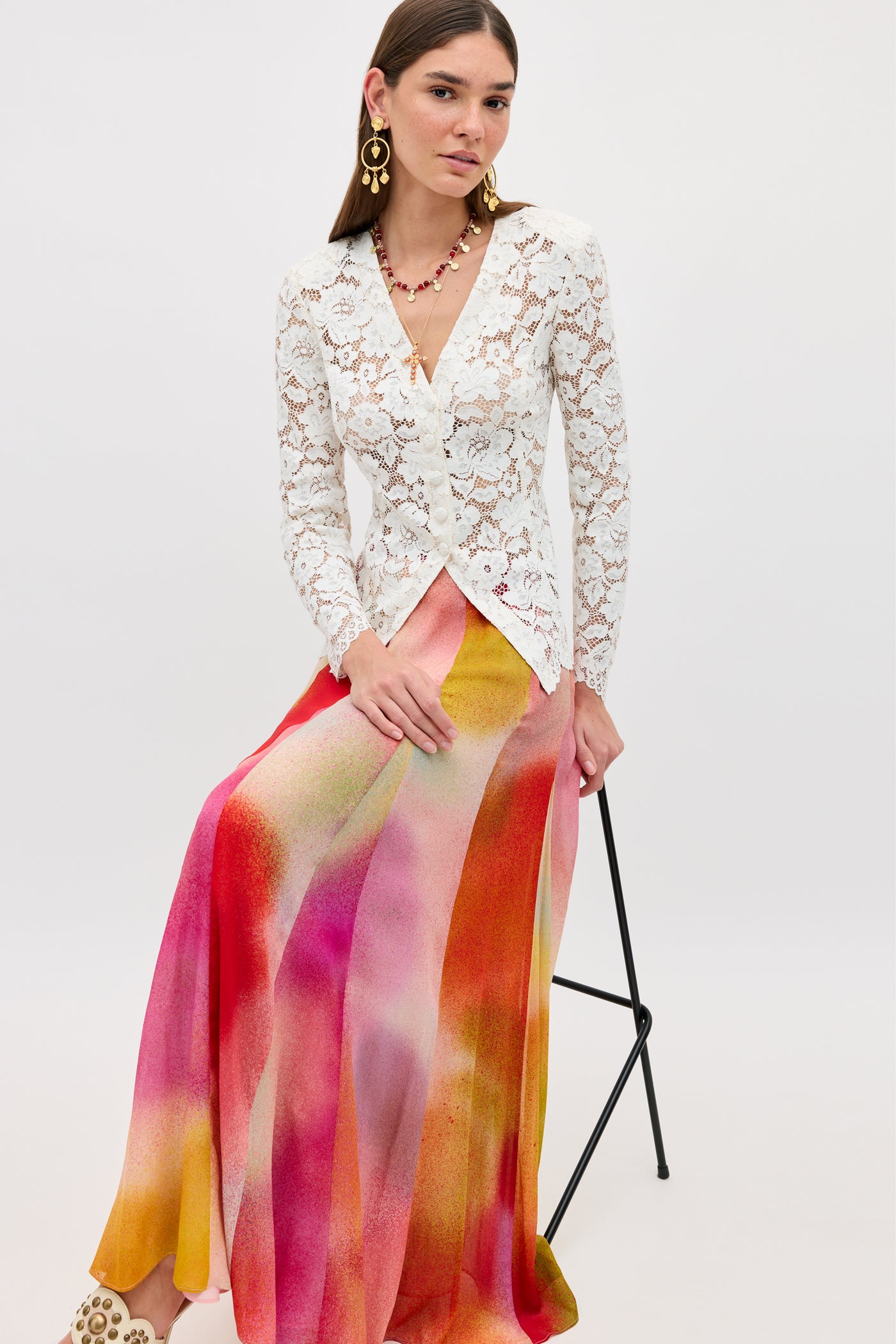 A woman sits on a stool wearing a white lace long-sleeve top and the Billy - Jessica Alexandra Multi maxi skirt with vibrant pink, red, and yellow Liberty stripes, accessorized with gold earrings, a necklace, and studded shoes—ideal for spring/summer.