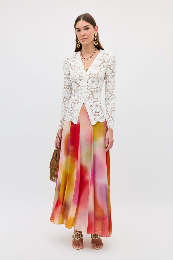 A woman wears the Billy - Jessica Alexandra Multi, a flowing Liberty fabric midi skirt with pink, orange, and yellow patterns, paired with a white lace long-sleeve top—perfect for spring/summer. She holds a brown woven handbag and wears statement earrings and sandals.