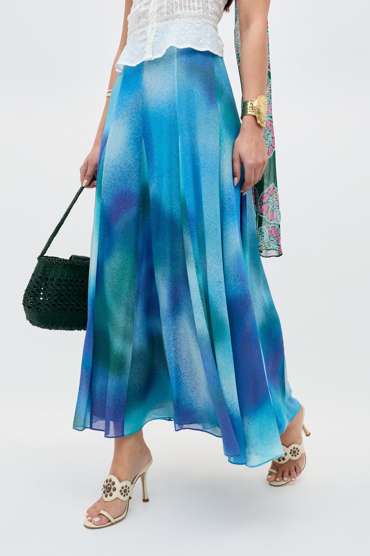 Dressed in the "Billy - Jessica Alexandra Blue" midi skirt—crafted from luxurious Liberty fabric with a graceful blue and purple gradient—a woman pairs it with a crisp white sleeveless top, cream heeled sandals featuring delicate floral cutouts, a dark green woven handbag, and gold jewelry. This effortlessly chic ensemble is perfect for spring or summer days, set against a light background.