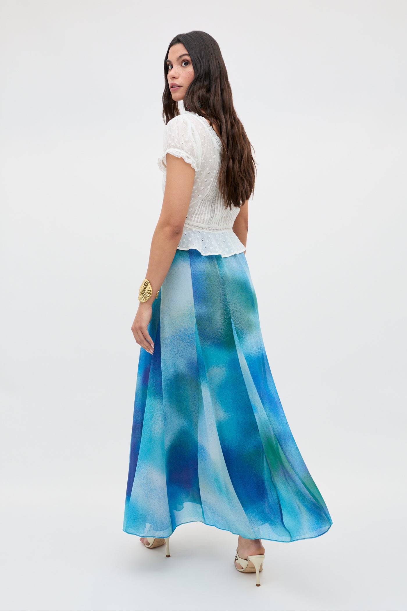 Woman with long dark hair wearing a white short-sleeve blouse and the Jessica Alexandra Blue "Billy" maxi skirt, crafted from flowing Liberty fabric, standing in heels and looking over her shoulder against a plain background—perfect inspiration for effortless spring/summer dressing.