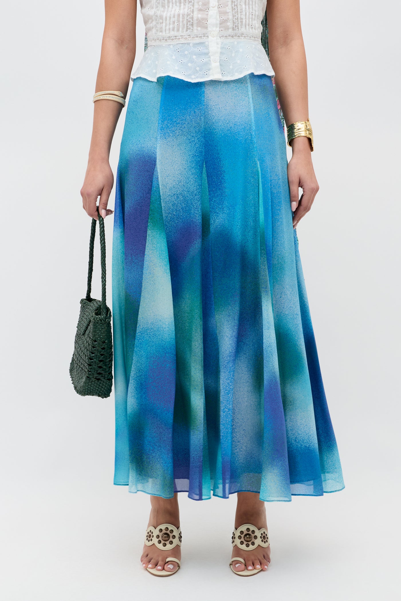 A woman wearing a white sleeveless top, the Billy - Jessica Alexandra Blue flowy blue and purple ombre midi skirt—perfect for spring or summer—beige cutout sandals, a gold bracelet, and holding a dark green woven handbag.