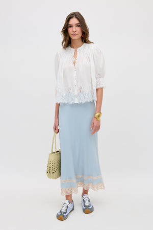 A woman with loose hair and a neutral expression stands against a plain background, wearing the Costanza - Bluebell light blue maxi skirt with lace trim, a white blouse with lace details, gray sneakers, and holding a woven straw bag.