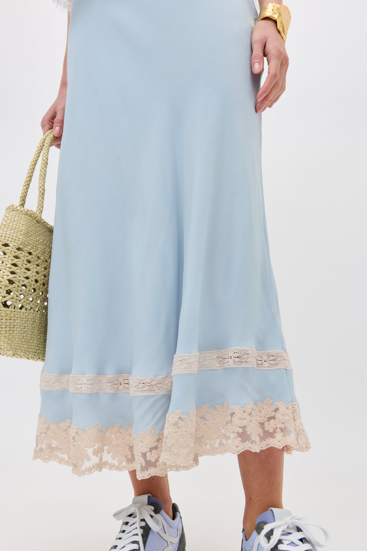 A person wearing the Costanza - Bluebell light blue skirt with beige lace trim, gray sneakers, a gold bracelet, and holding a woven straw bag. The photo is cropped at the torso.