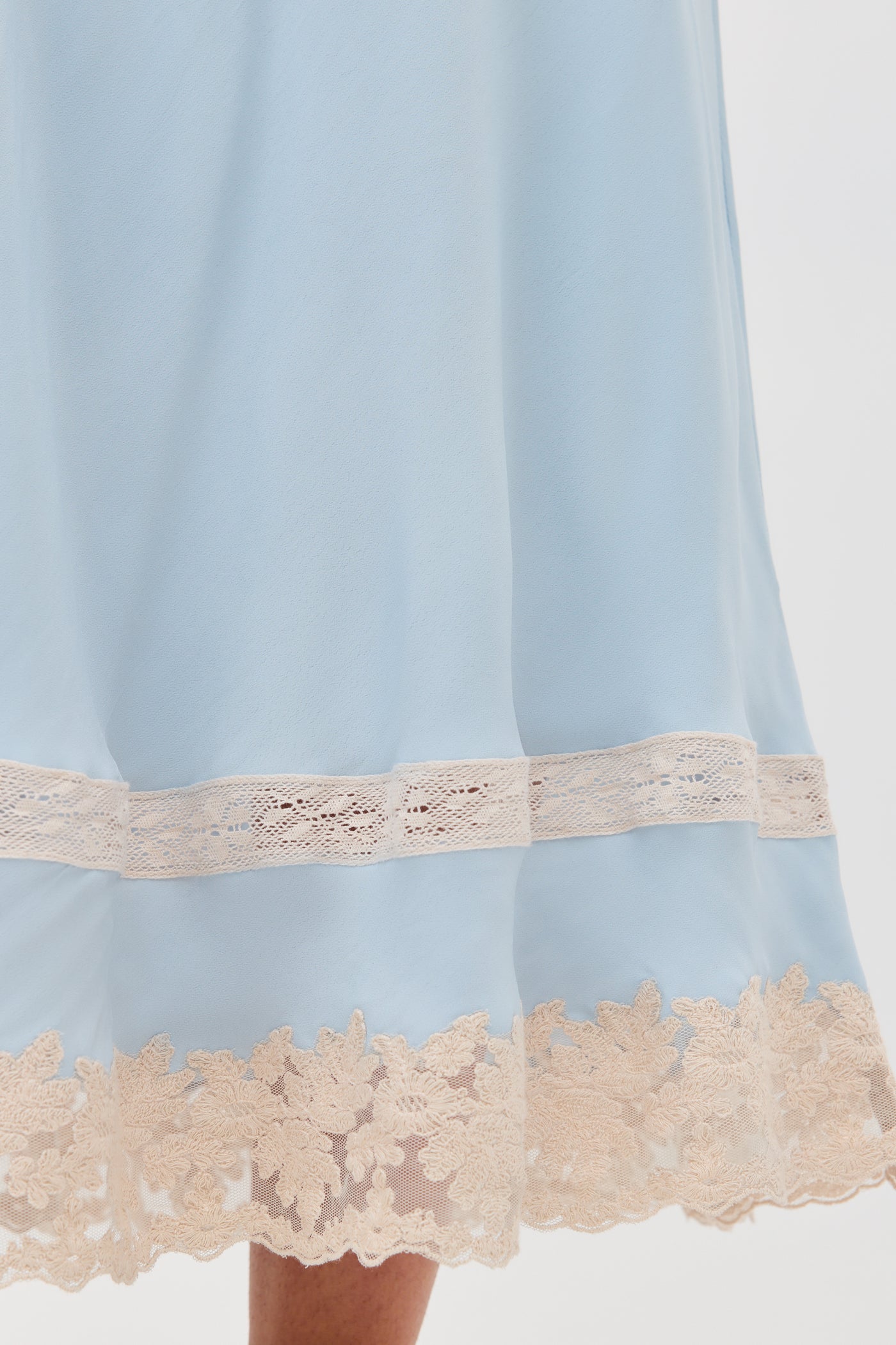 A close-up of the Costanza - Bluebell dress shows soft, light blue fabric with a cream lace trim at the hem and a horizontal lace detail above it, creating an elegant, flowing look.