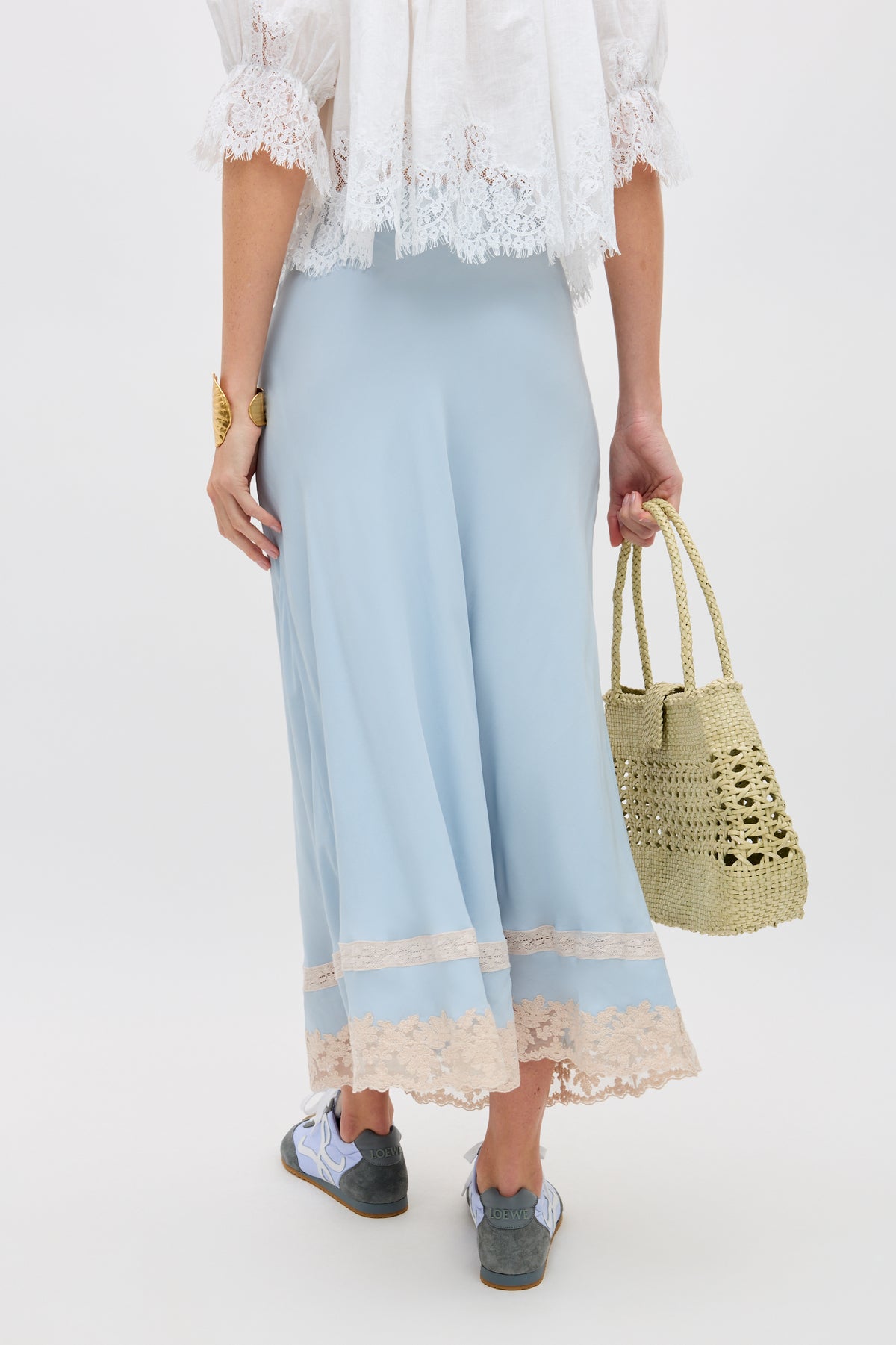 A person wearing the Costanza - Bluebell light blue skirt with beige lace trim, a white lace top, and sneakers holds a woven straw bag. Photo is taken from behind against a plain white background.