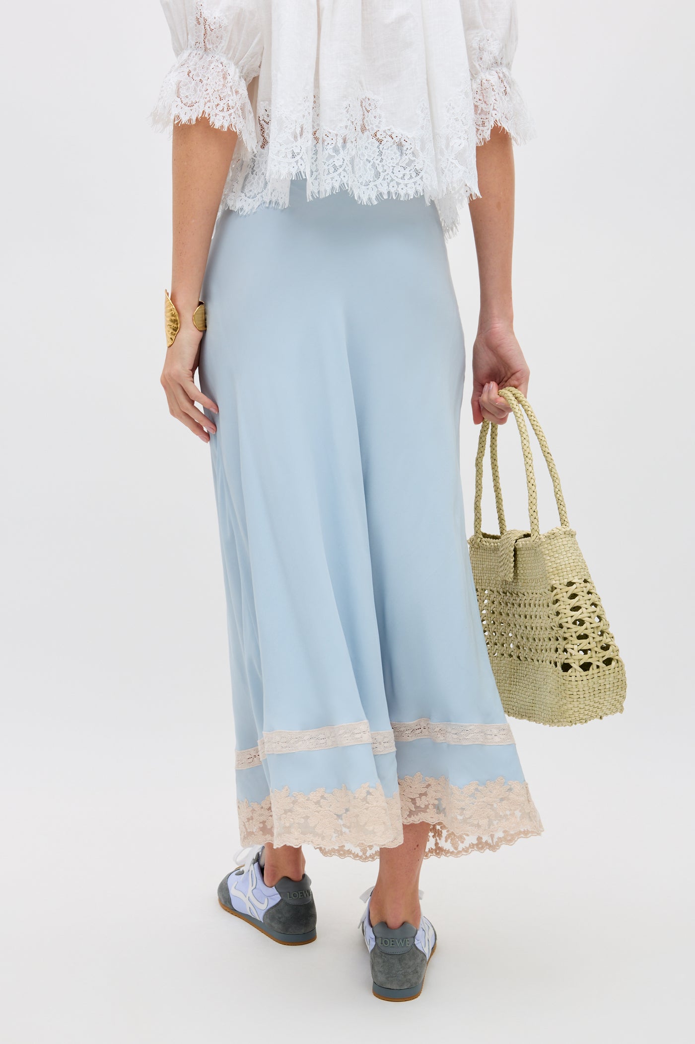 A person wearing the Costanza - Bluebell light blue skirt with beige lace trim, a white lace top, and sneakers holds a woven straw bag. Photo is taken from behind against a plain white background.