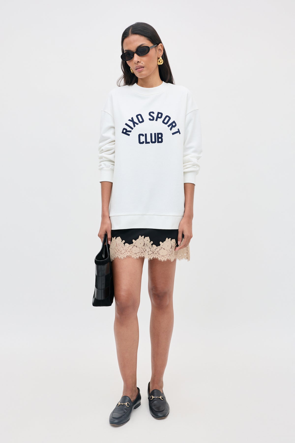 Woman wearing sunglasses, a white RIXO SPORT CLUB sweatshirt, a black skirt with beige lace trim, black loafers, and holding the Ariah - Black handbag, standing in front of a plain light background.