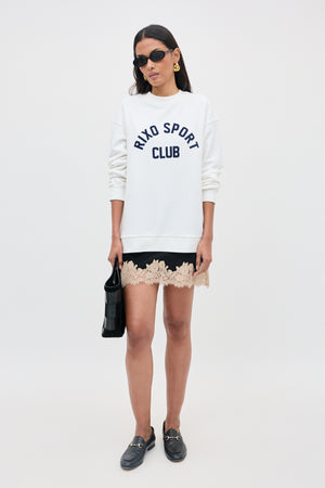 Woman wearing sunglasses, a white RIXO SPORT CLUB sweatshirt, a black skirt with beige lace trim, black loafers, and holding the Ariah - Black handbag, standing in front of a plain light background.