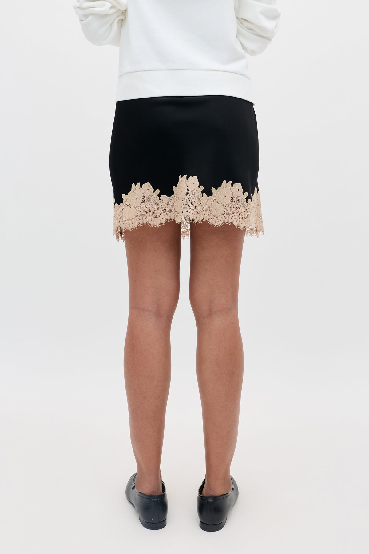 A person standing with their back to the camera, wearing the Ariah - Black mini skirt featuring beige lace trim, paired with a white top and black shoes.