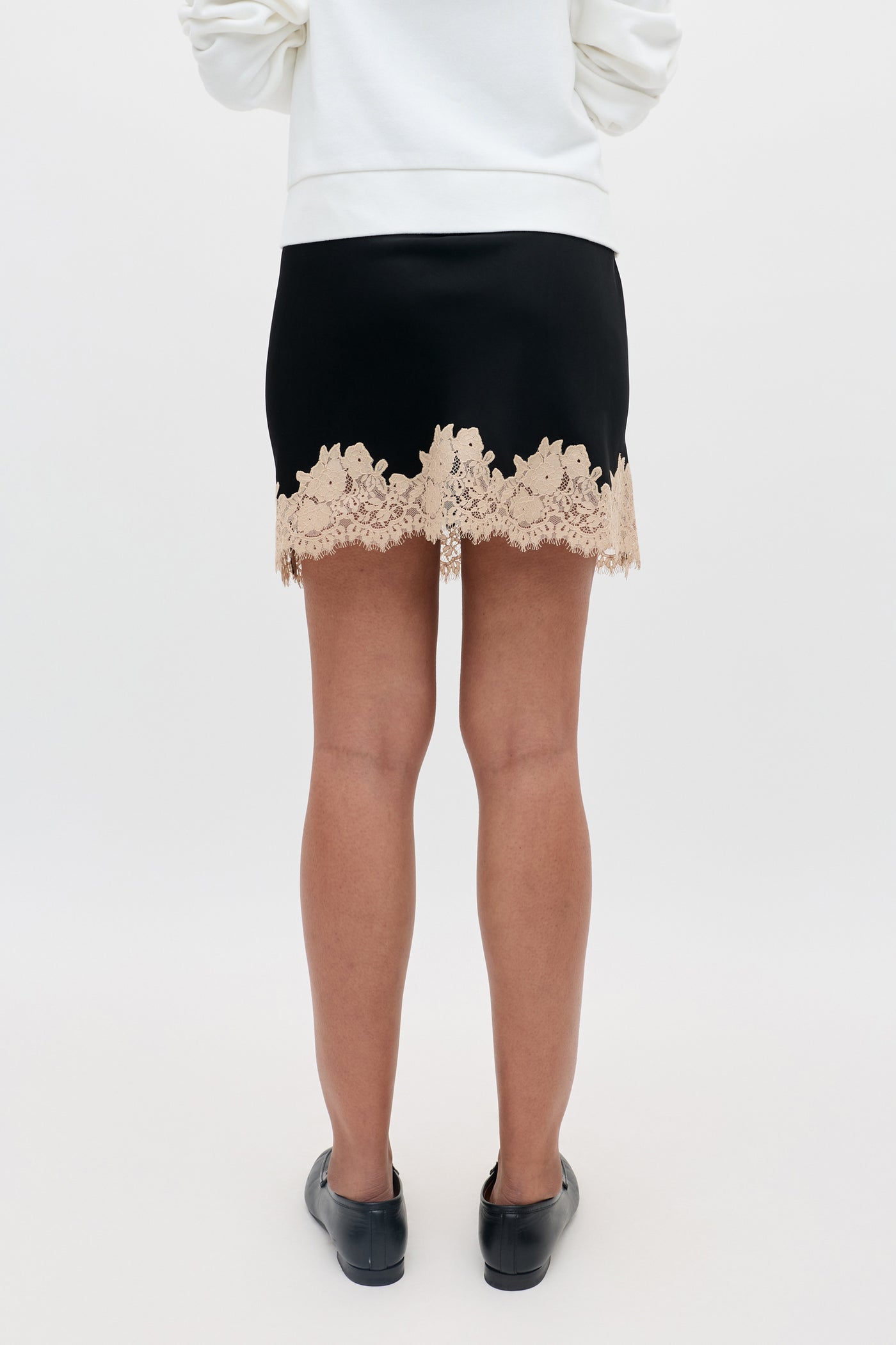A person standing with their back to the camera, wearing the Ariah - Black mini skirt featuring beige lace trim, paired with a white top and black shoes.