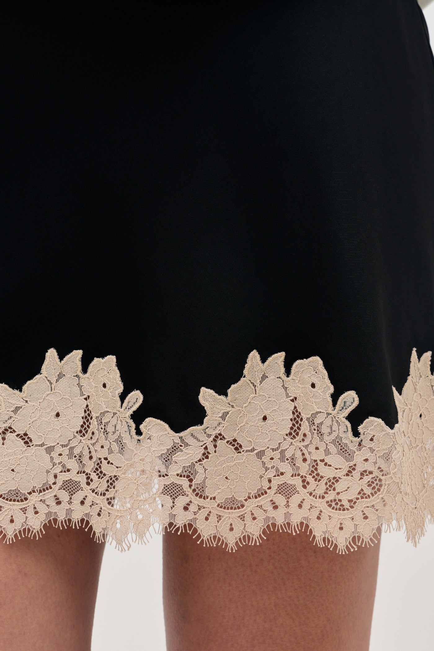 Close-up of the hem of Ariah - Black, a black garment featuring beige floral lace trim, worn by a person with partially visible legs.