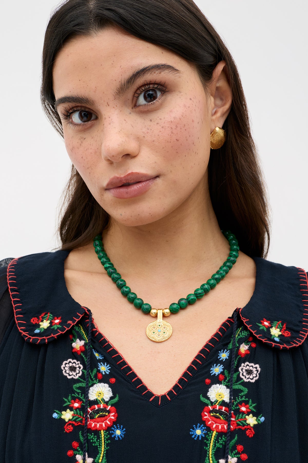 A woman with long dark hair, in a navy dress with colorful floral embroidery and gold earrings, wears the Ischia - Green beaded necklace with a golden pendant, looking directly at the camera against a light background.