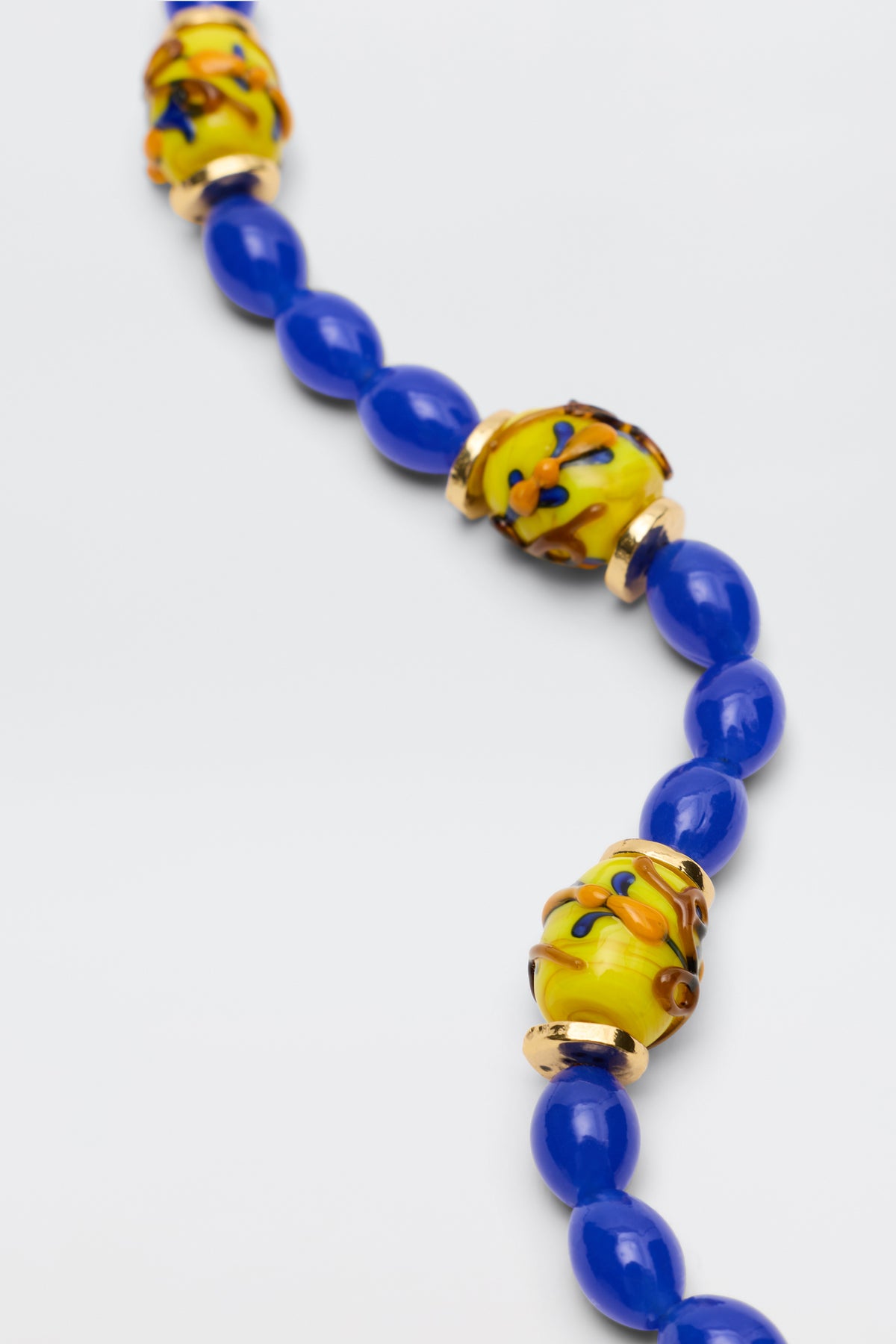 A beaded Denali - Multi necklace featuring bright blue oval beads and three larger yellow beads with blue and brown designs, spaced along the strand with delicate gold-plated beads, shown against a plain white background.