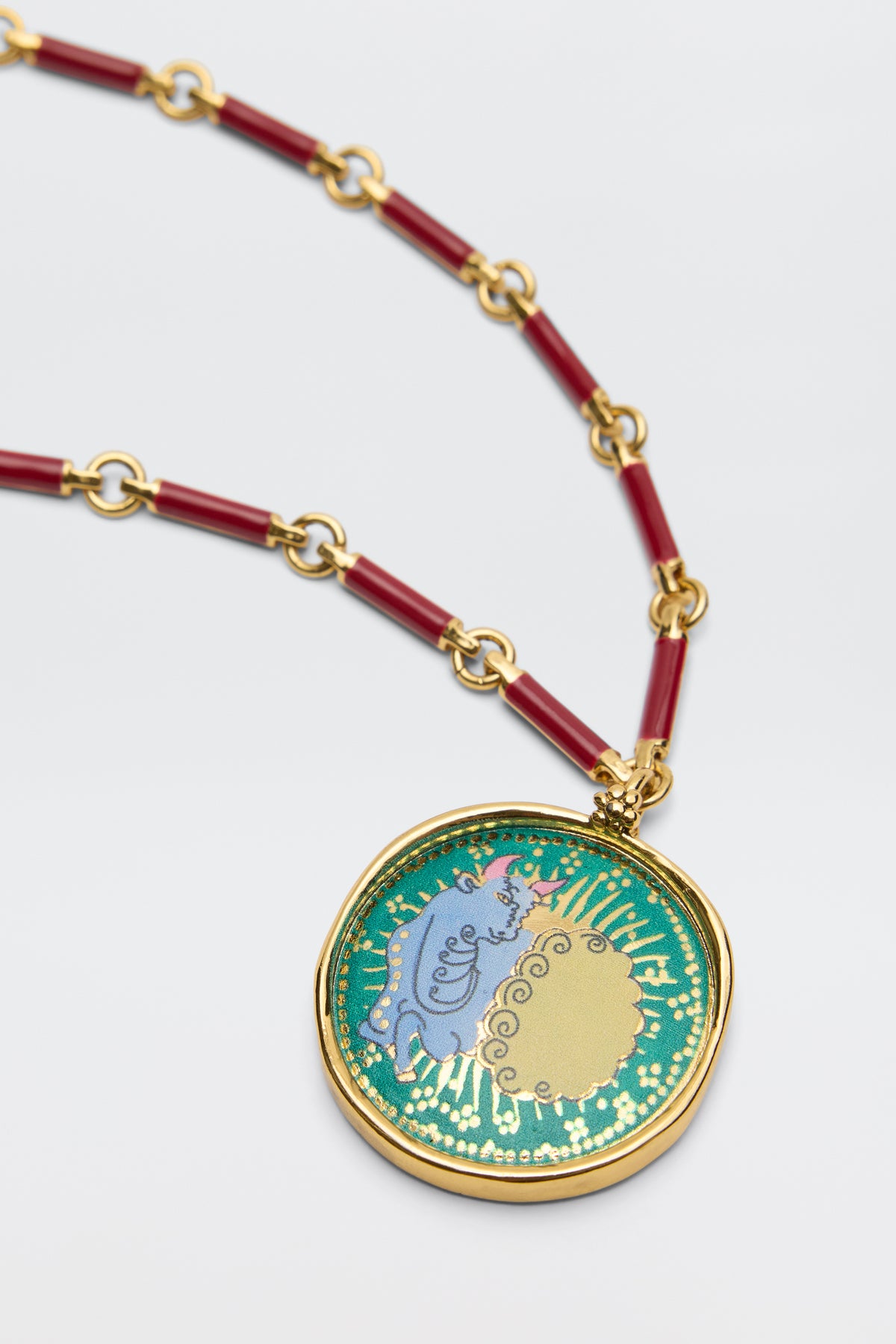 The Ari - Astro Taurus necklace features a gold link chain with red segments and a round pendant showcasing a blue elephant, gold sun, and green background accented with decorative gold details.