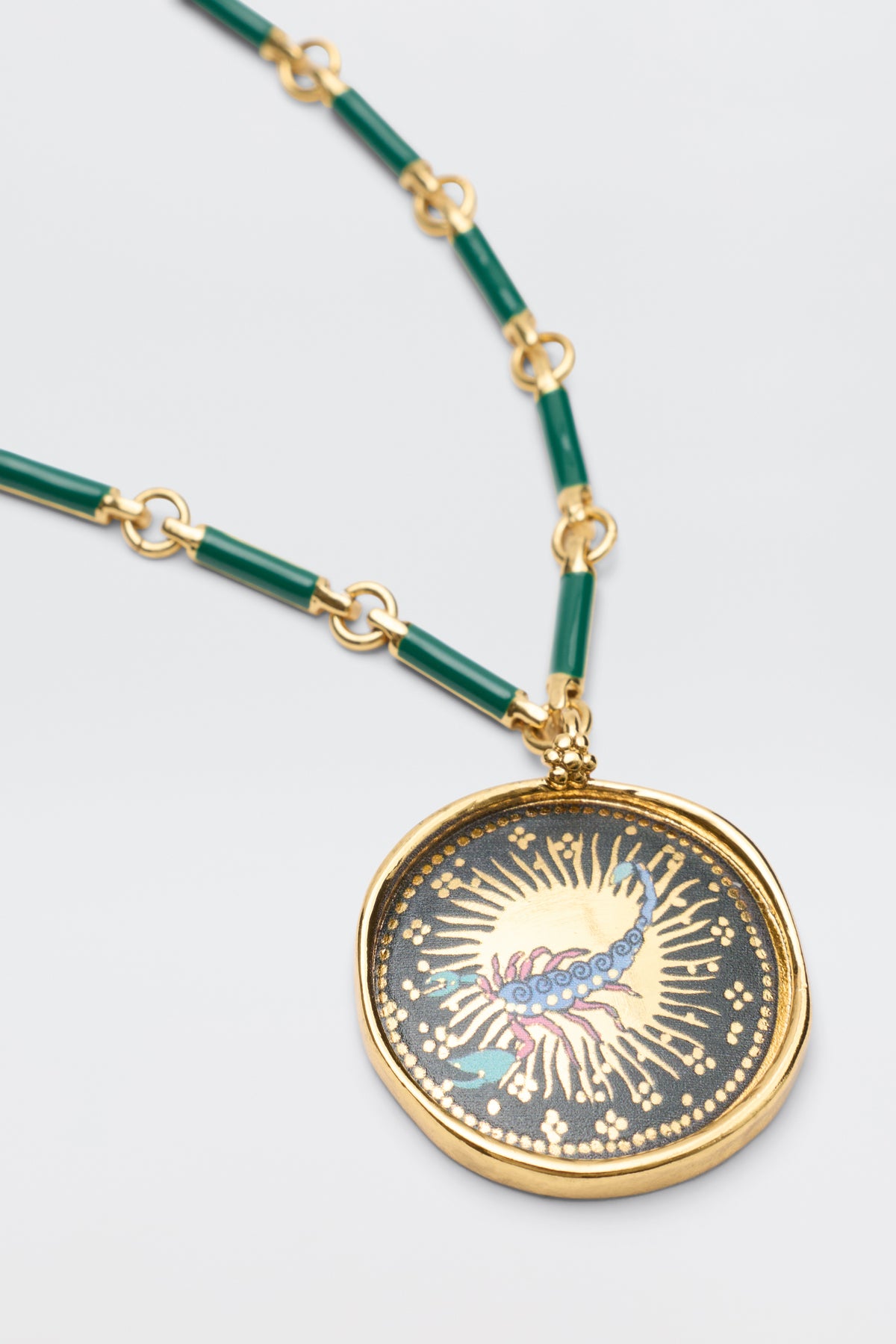 The Ari - Astro Scorpio necklace features green cylindrical beads and a bold gold disc pendant with a colorful abstract design, possibly a centipede, set against a dark background with gold sunburst accents.