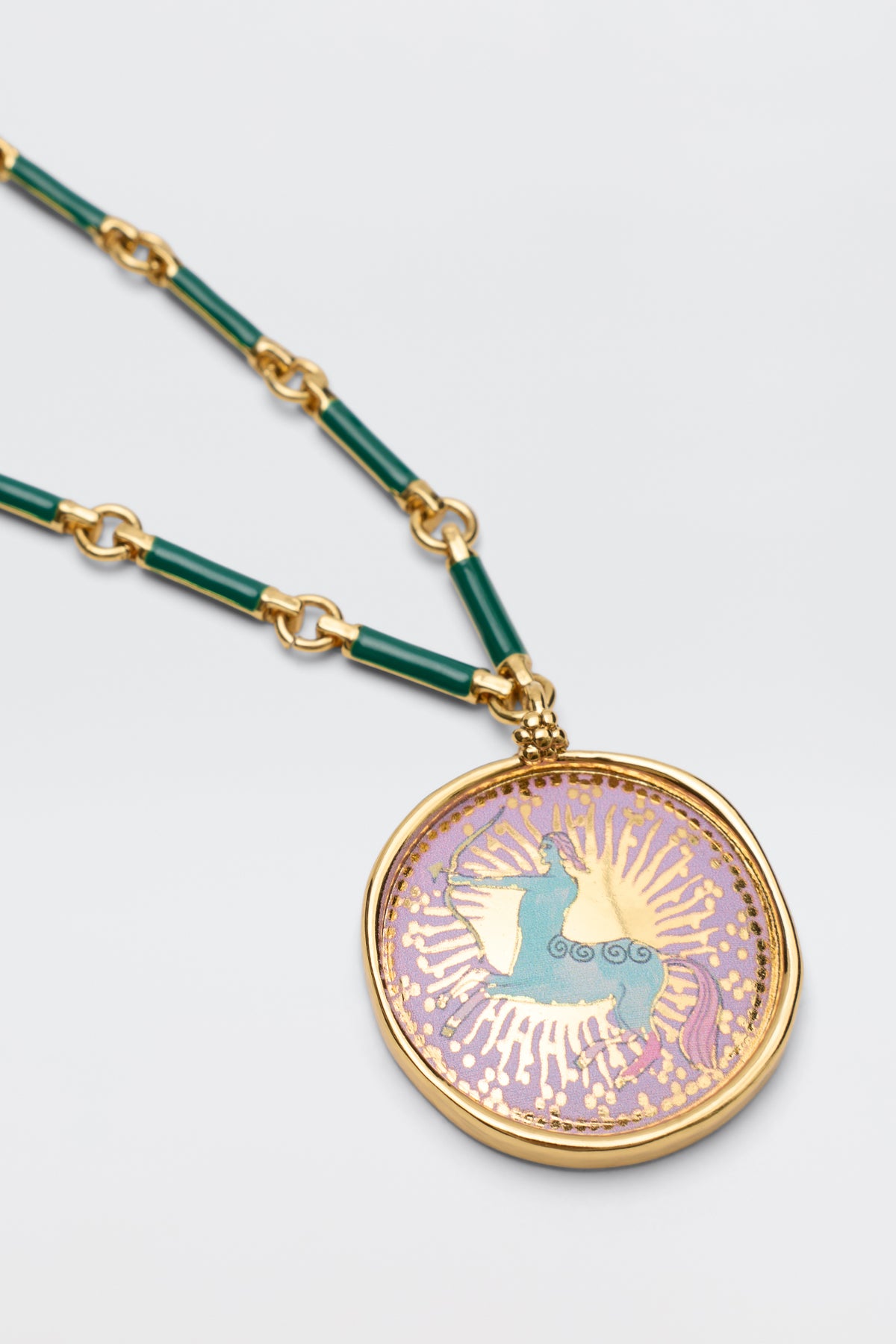 Ari - Astro Sagittarius: A gold zodiac necklace with green links and a round disc charm featuring a blue horse on a pink and gold sunburst background.