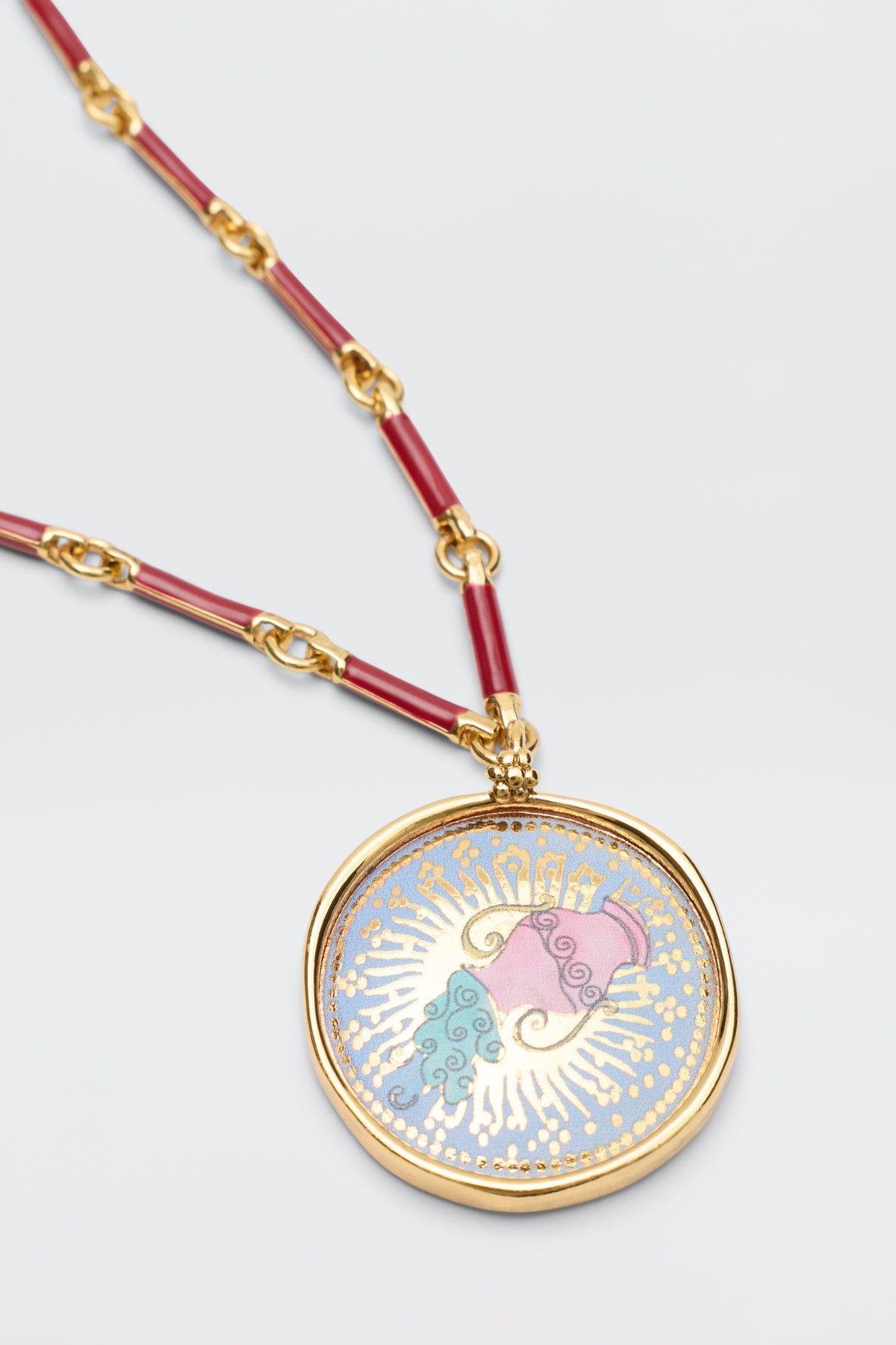 Ari - Astro Aquarius: A gold necklace with a bold disc charm featuring red segments, plus a round pendant of a pink and blue swirling heart with yellow rays on a light blue background.