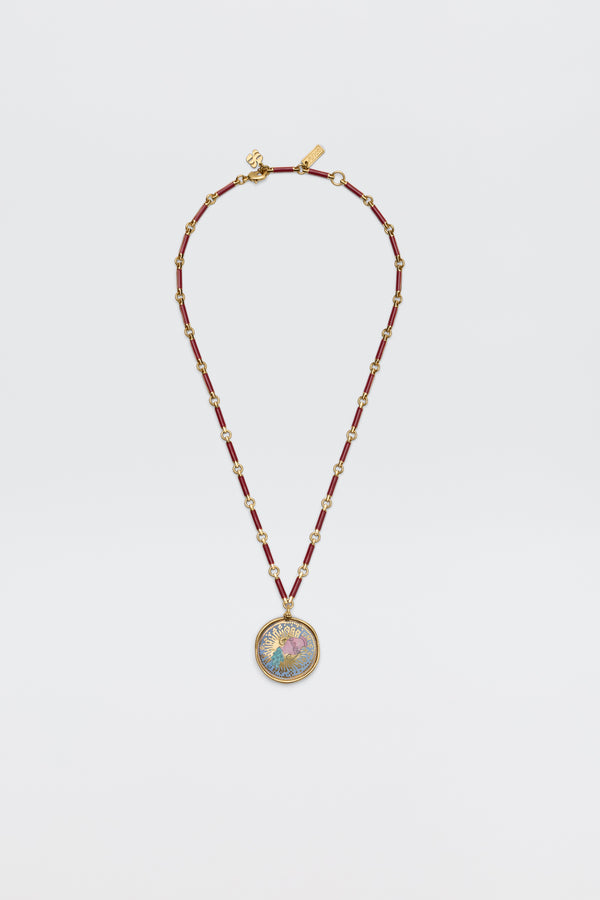 The Ari - Astro Aquarius necklace features gold zodiac detailing, red cylindrical beads, small gold spacers, and a statement disc charm with a colorful, intricate design at the center. Displayed against a plain white background.