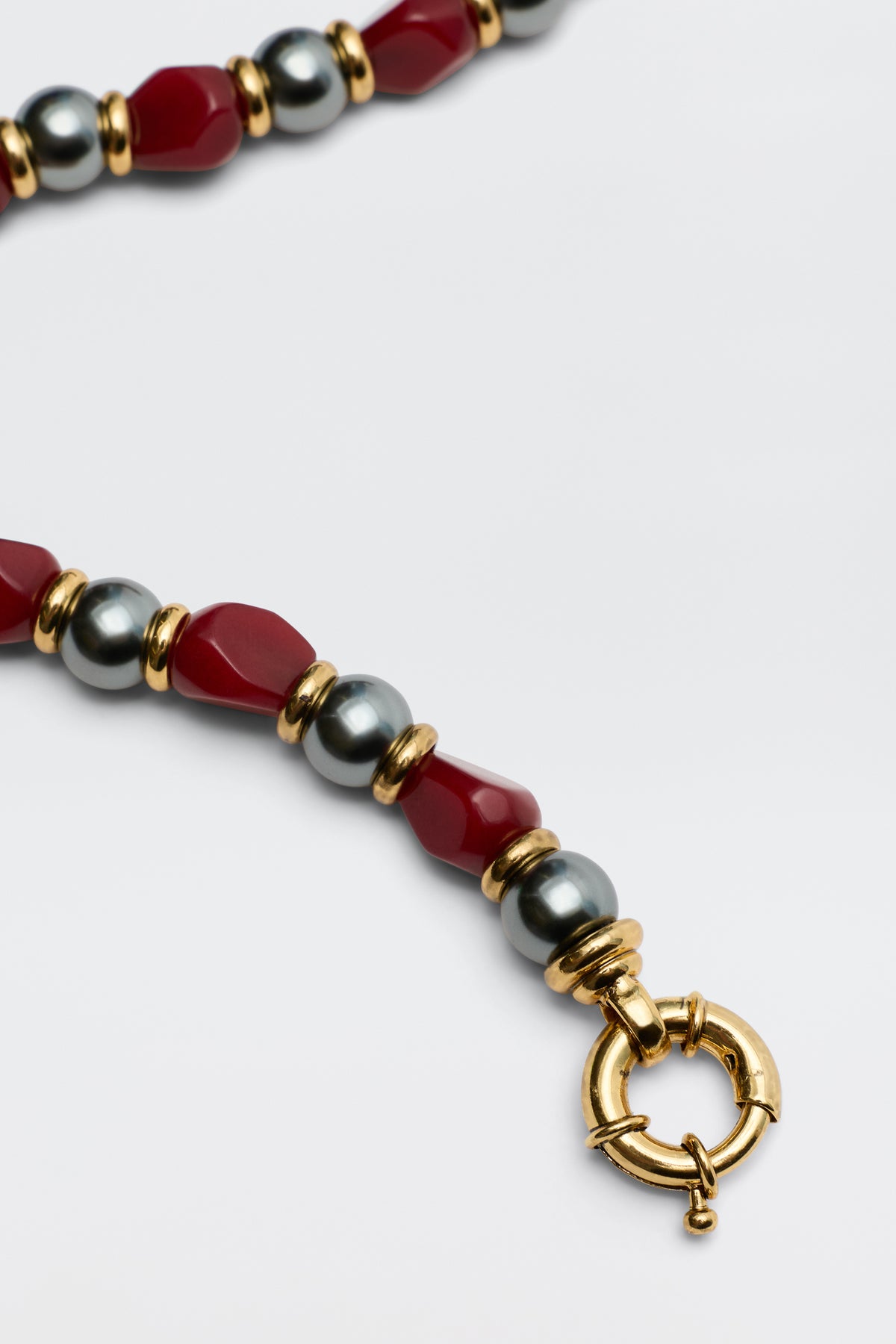 A close-up of the Palmara - Burgundy necklace, showcasing alternating red and metallic grey beads with gold accents and a round gold clasp on a plain white background.