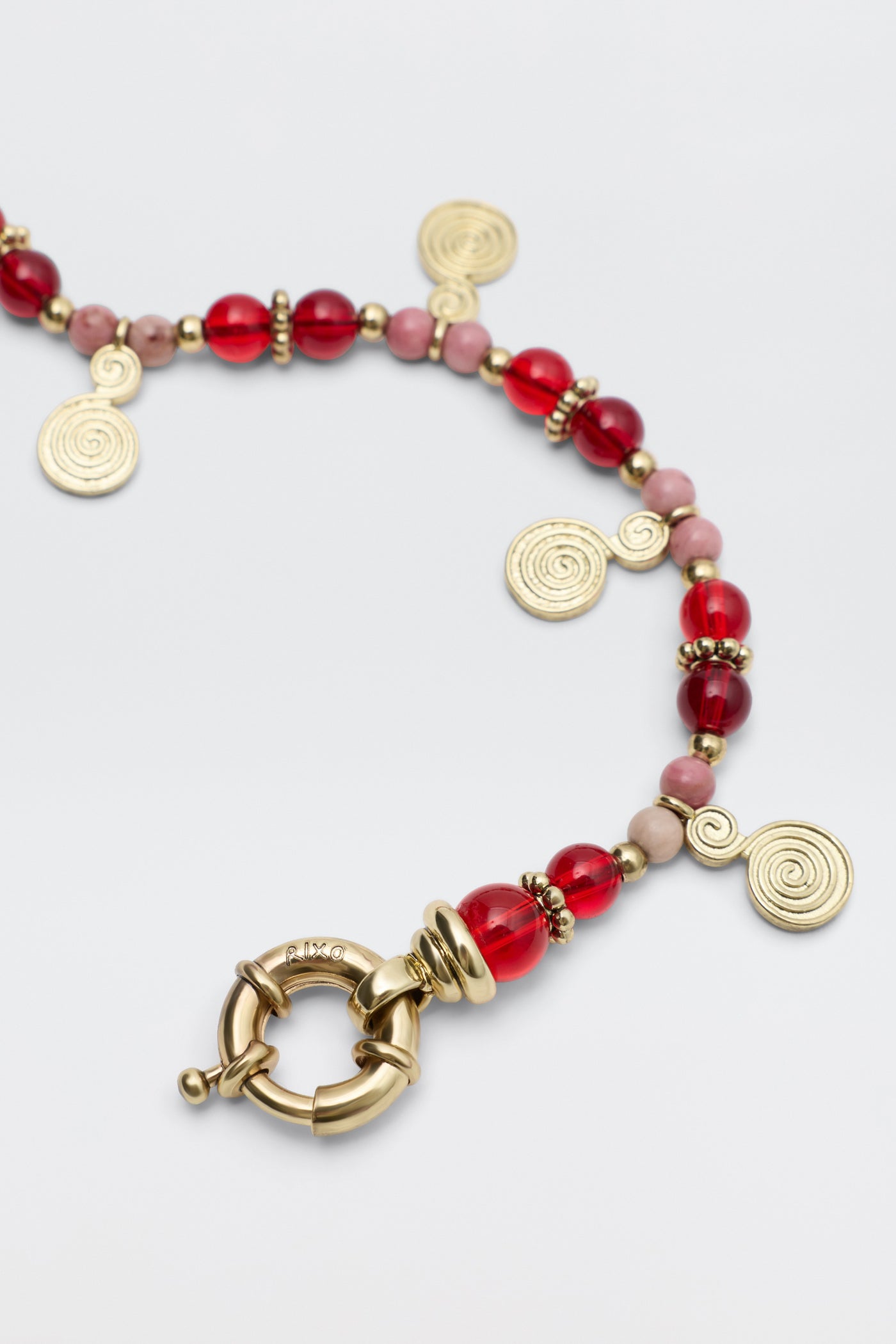 Tala - Burgundy: A beaded necklace with burgundy and pink beads, gold spacer beads, gold spiral charms, and a large round gold clasp, displayed on a light background.