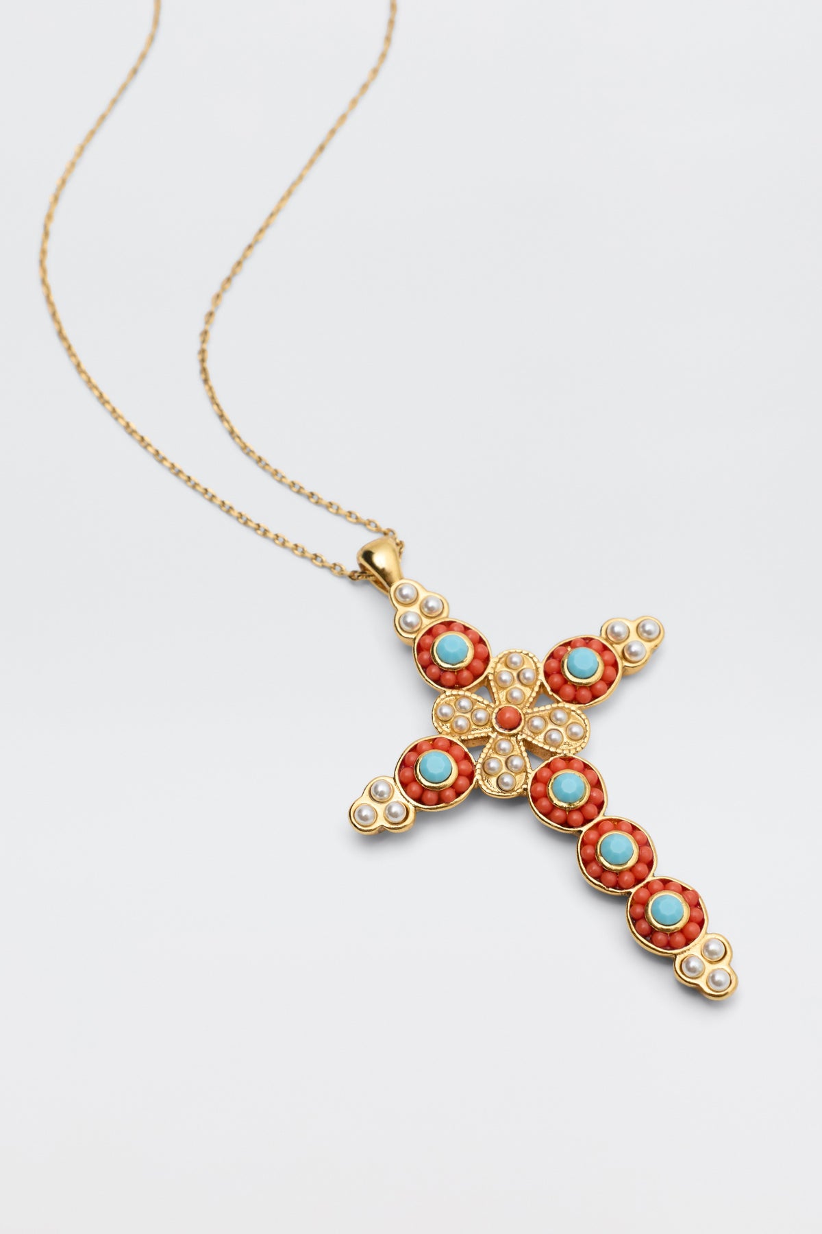 The Lenka - Gold is a gold chain necklace with an ornate cross pendant, featuring red coral and turquoise stones in intricate patterns, accented by small white pearls. Set against a plain, light-colored background.
