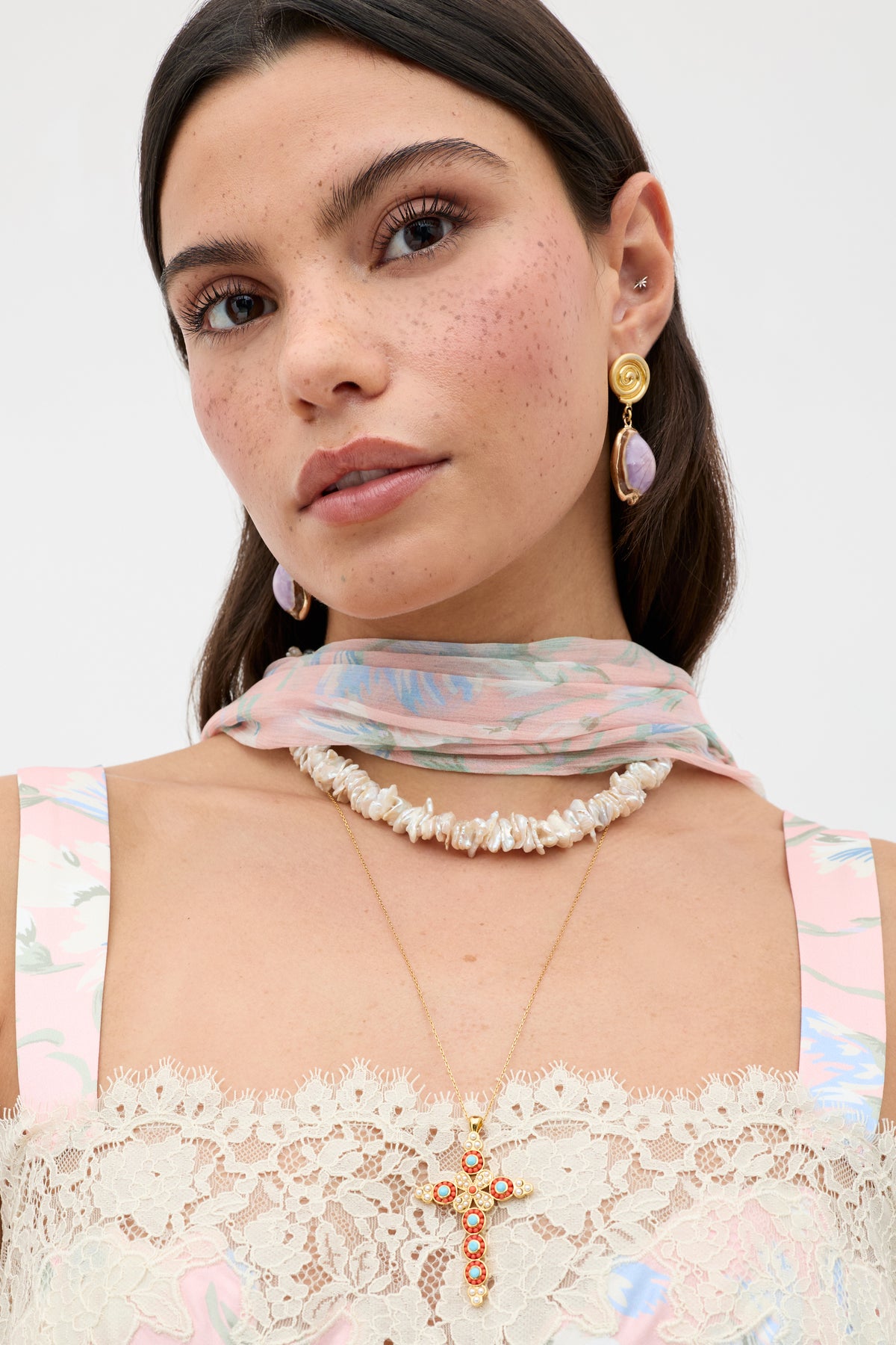 A woman with long brown hair wears the Lenka - Gold earrings featuring purple stones, a pastel floral scarf, pearl necklace, gold cross pendant, natural makeup, and a lace-trimmed floral dress.