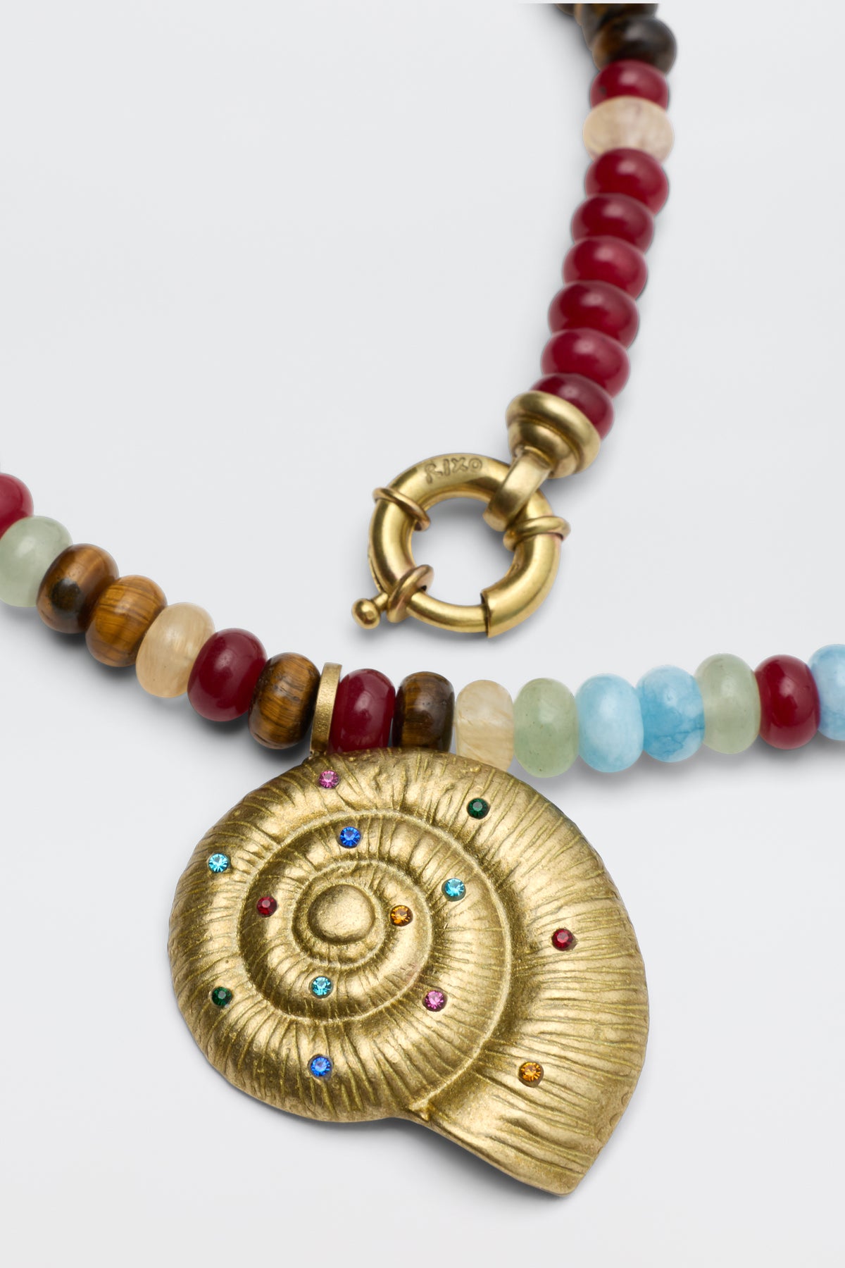 The Parma - Multi is a beaded necklace with red, blue, brown, and cream stones, featuring a gold spiral shell pendant decorated with small multicolored gemstones.
