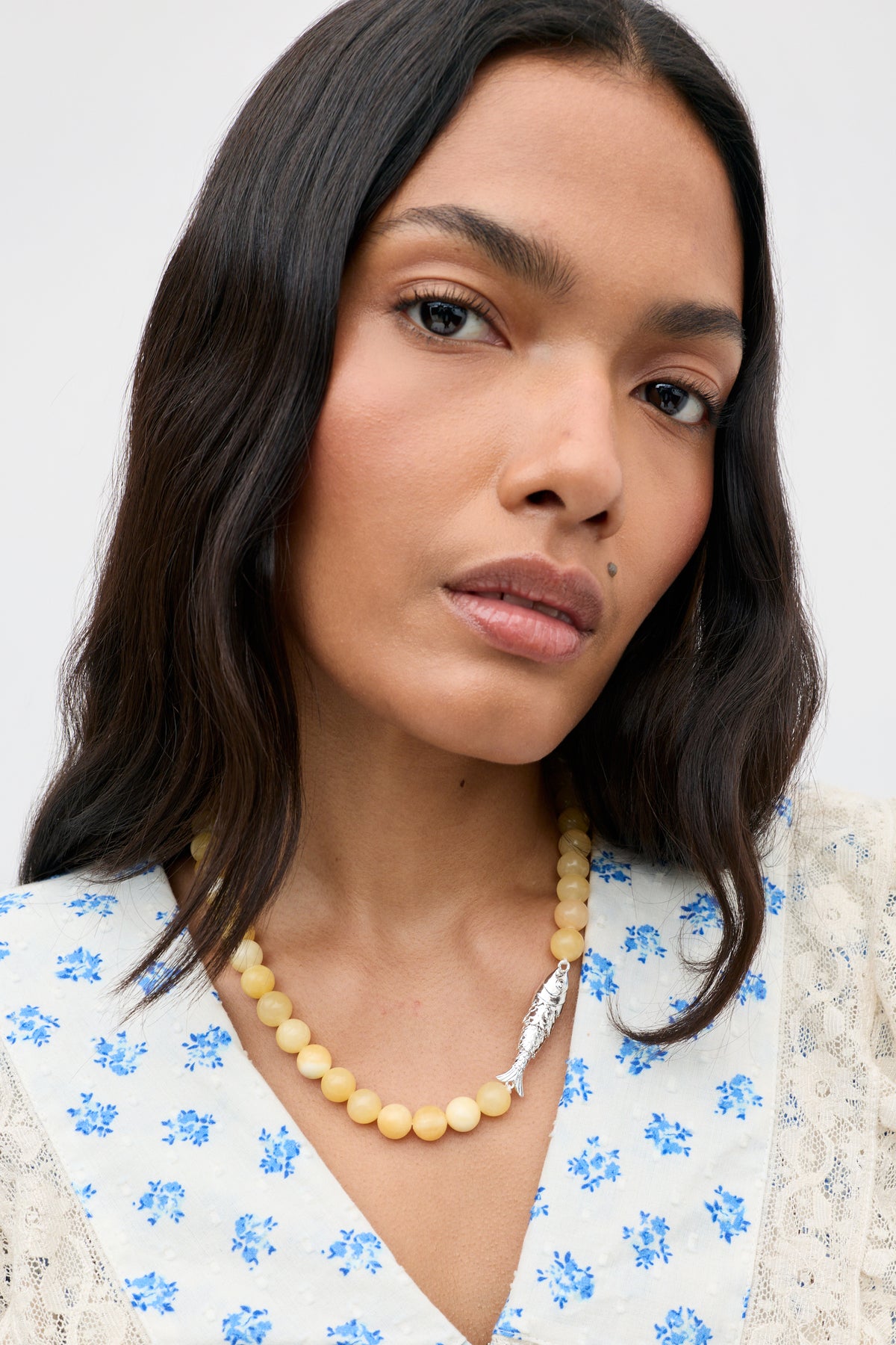 A woman with long dark hair wears the Amoura - Lemon cream beaded necklace featuring a silver feather pendant, paired with a white blouse adorned with blue floral prints. She looks directly at the camera against a plain background.