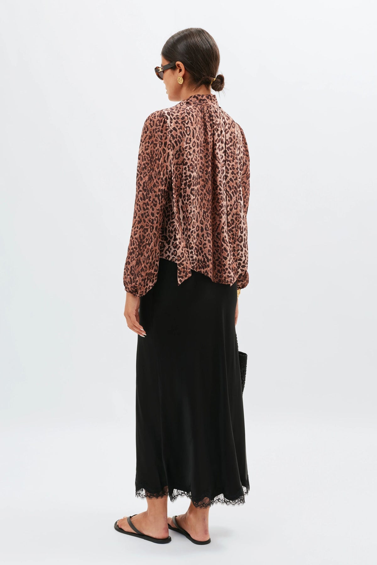 A woman stands facing away, wearing the Moss - Leopard Brown blouse with a black lace-trim midi skirt, black sandals, sunglasses, and a small black bag. Her hair is styled in a low bun.