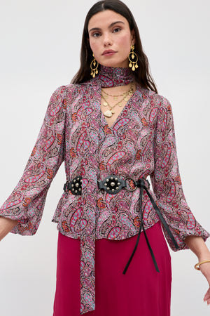 A woman wearing the Moss - Bourton Red long-sleeved vintage-inspired paisley blouse with matching neck scarf, bold earrings, layered necklaces, a black belt with floral accents, and a magenta skirt, standing against a plain light background.