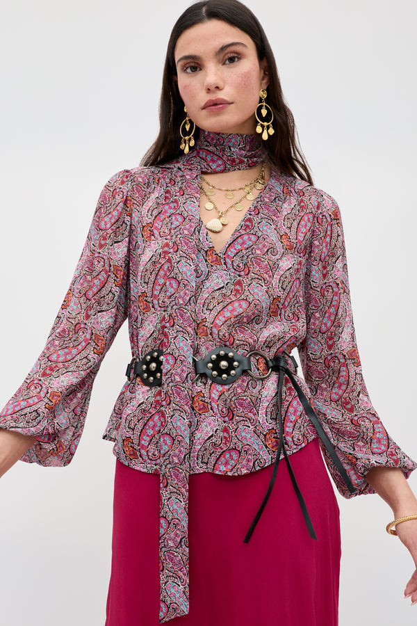 A woman wearing the Moss - Bourton Red long-sleeved vintage-inspired paisley blouse with matching neck scarf, bold earrings, layered necklaces, a black belt with floral accents, and a magenta skirt, standing against a plain light background.