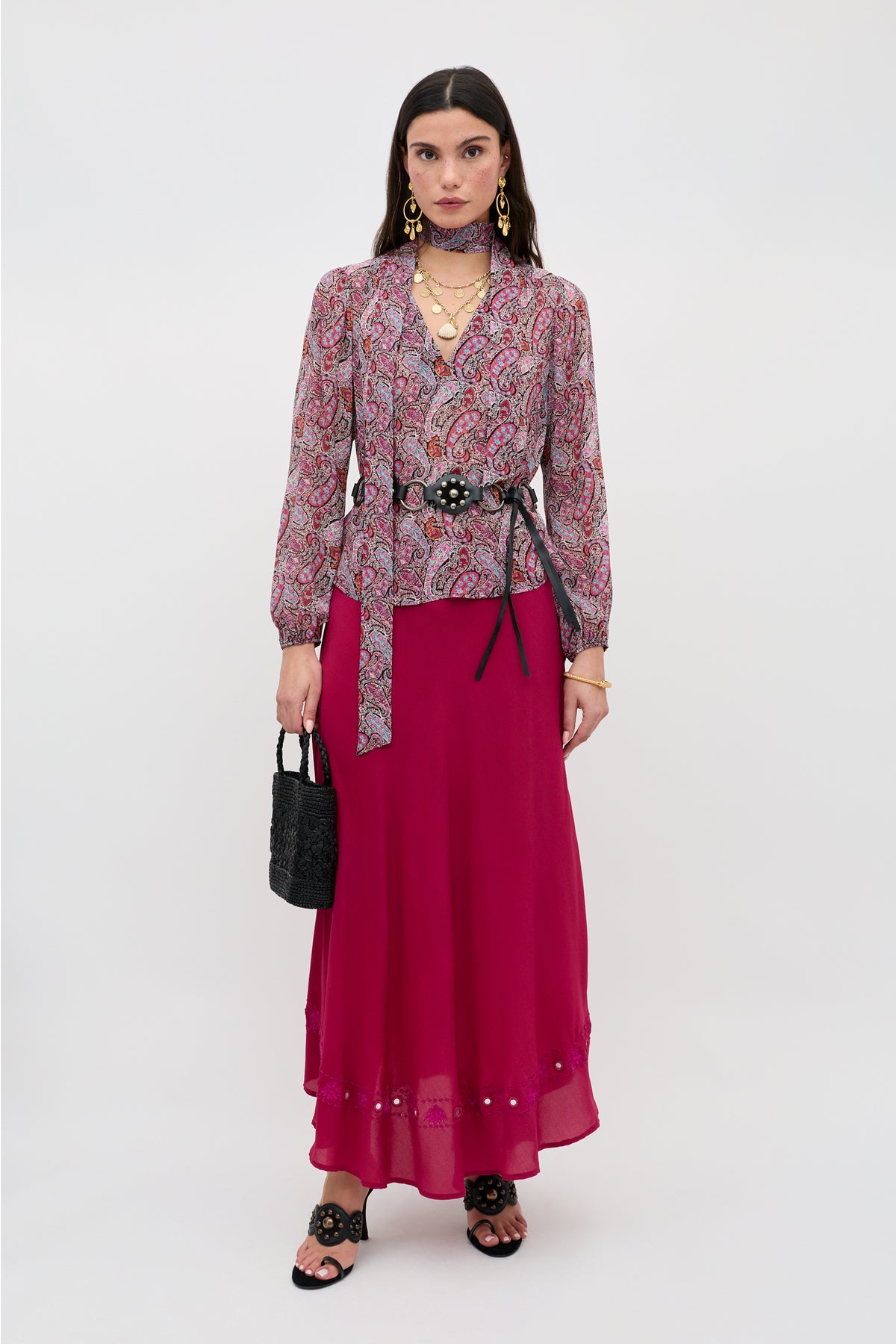 A woman wears the Moss - Bourton Red vintage-inspired blouse, paired with a magenta maxi skirt, black belt, and sandals. She accessorizes with a black handbag and layered gold jewelry, standing against a plain white background.