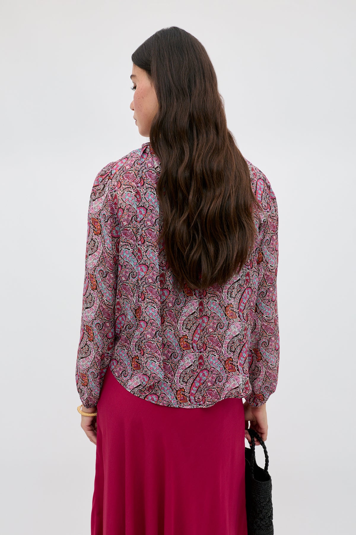 A person with long wavy brown hair, wearing a vintage-inspired blouse and magenta skirt, stands facing away holding the Moss - Bourton Red bag against a plain, light background.