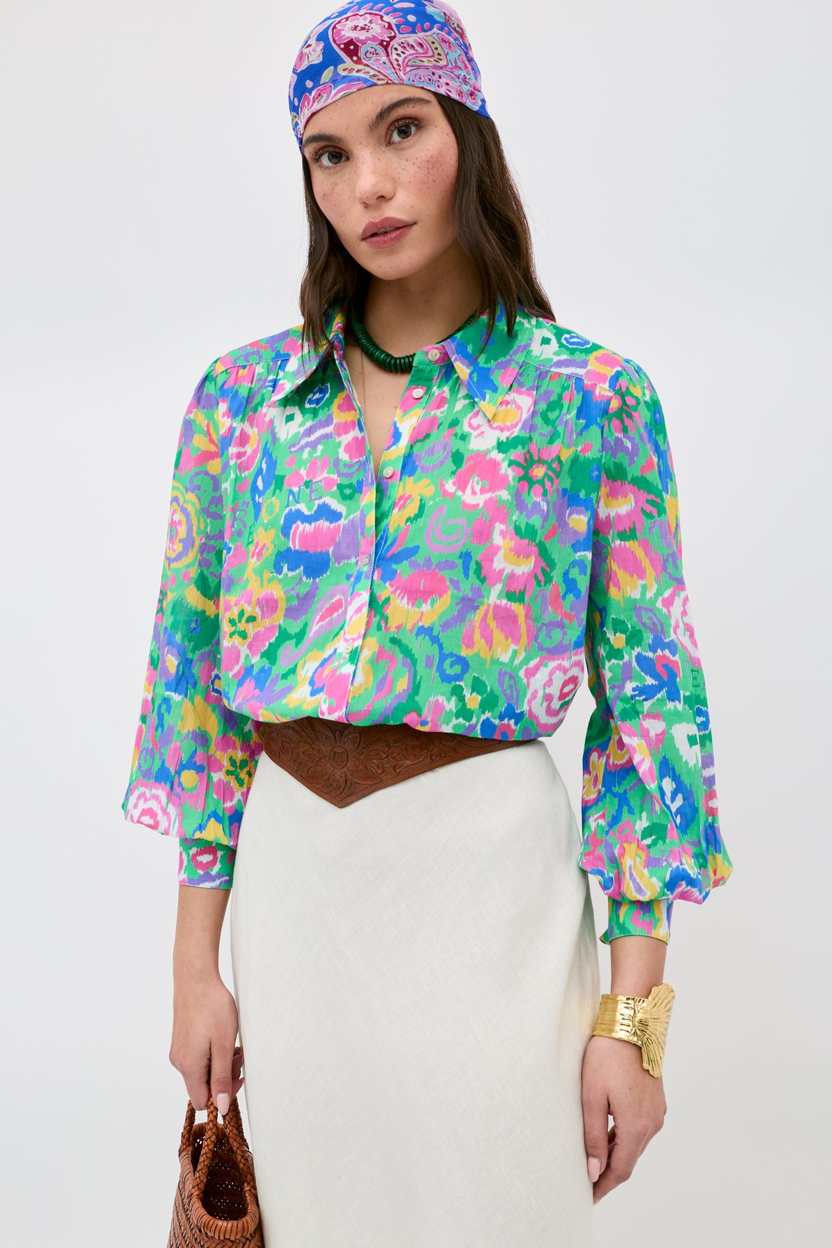 A woman stands against a plain background, wearing the Blake - Ikat Bloom Green floral blouse paired with a white skirt, patterned headscarf, brown belt, gold bracelet, green necklace, and holding a brown bag.