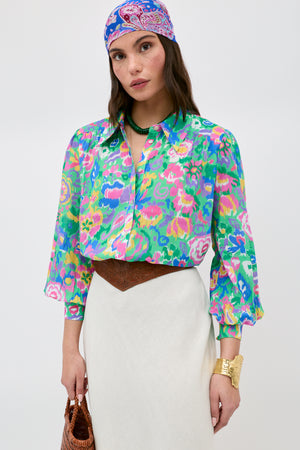 A woman stands against a plain background, wearing the Blake - Ikat Bloom Green floral blouse paired with a white skirt, patterned headscarf, brown belt, gold bracelet, green necklace, and holding a brown bag.