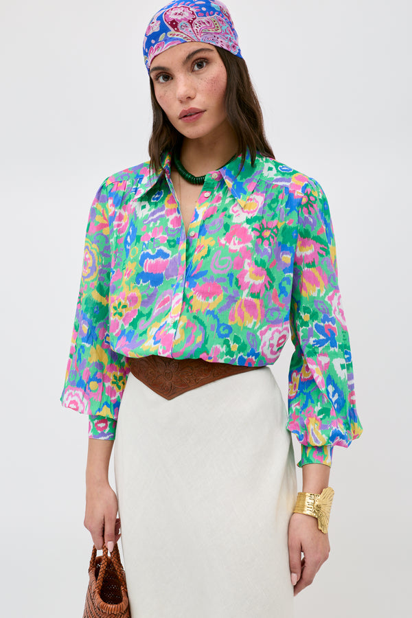 A woman stands against a plain background, wearing the Blake - Ikat Bloom Green floral blouse paired with a white skirt, patterned headscarf, brown belt, gold bracelet, green necklace, and holding a brown bag.