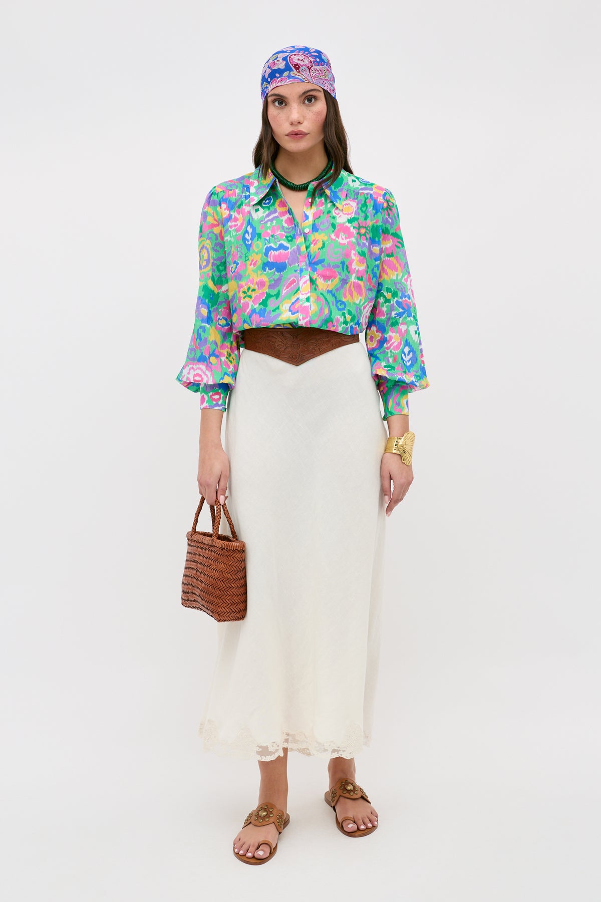 A woman stands against a plain white background wearing the Blake - Ikat Bloom Green shirt, styled with a white maxi skirt, brown belt, and sandals. She accessorizes with a vibrant headscarf, gold bracelet, and carries a woven bag.
