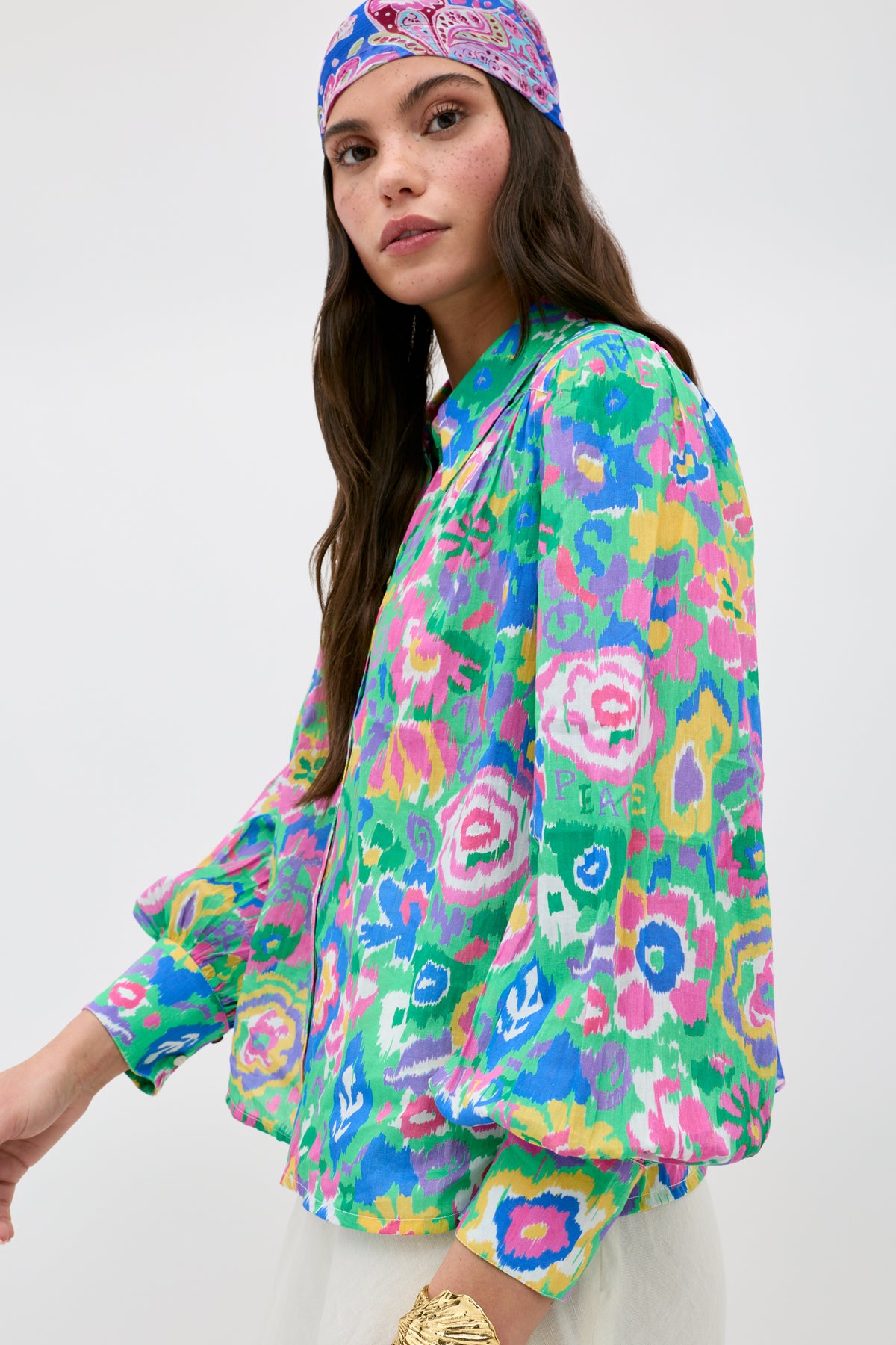 A woman wearing the "Blake - Ikat Bloom Green" shirt, featuring colorful pink, green, yellow, and blue patterns, paired with a matching headscarf and a gold bracelet, stands against a plain light background.