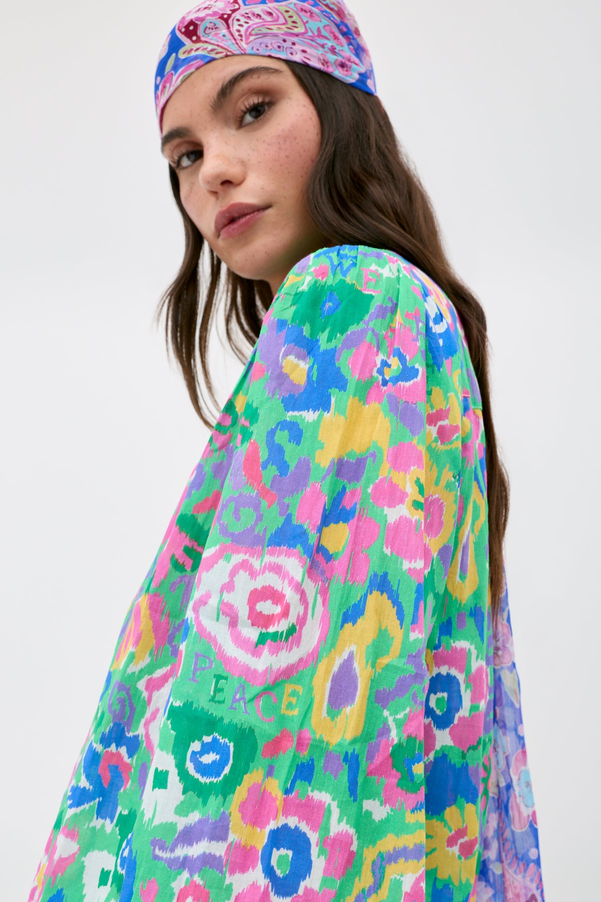 A woman wearing the Blake - Ikat Bloom Green dress and matching headscarf looks over her shoulder. The colorful, floral-patterned dress features bright pink, green, blue, and yellow hues, with "PEACE" and a peace symbol visible in the design.